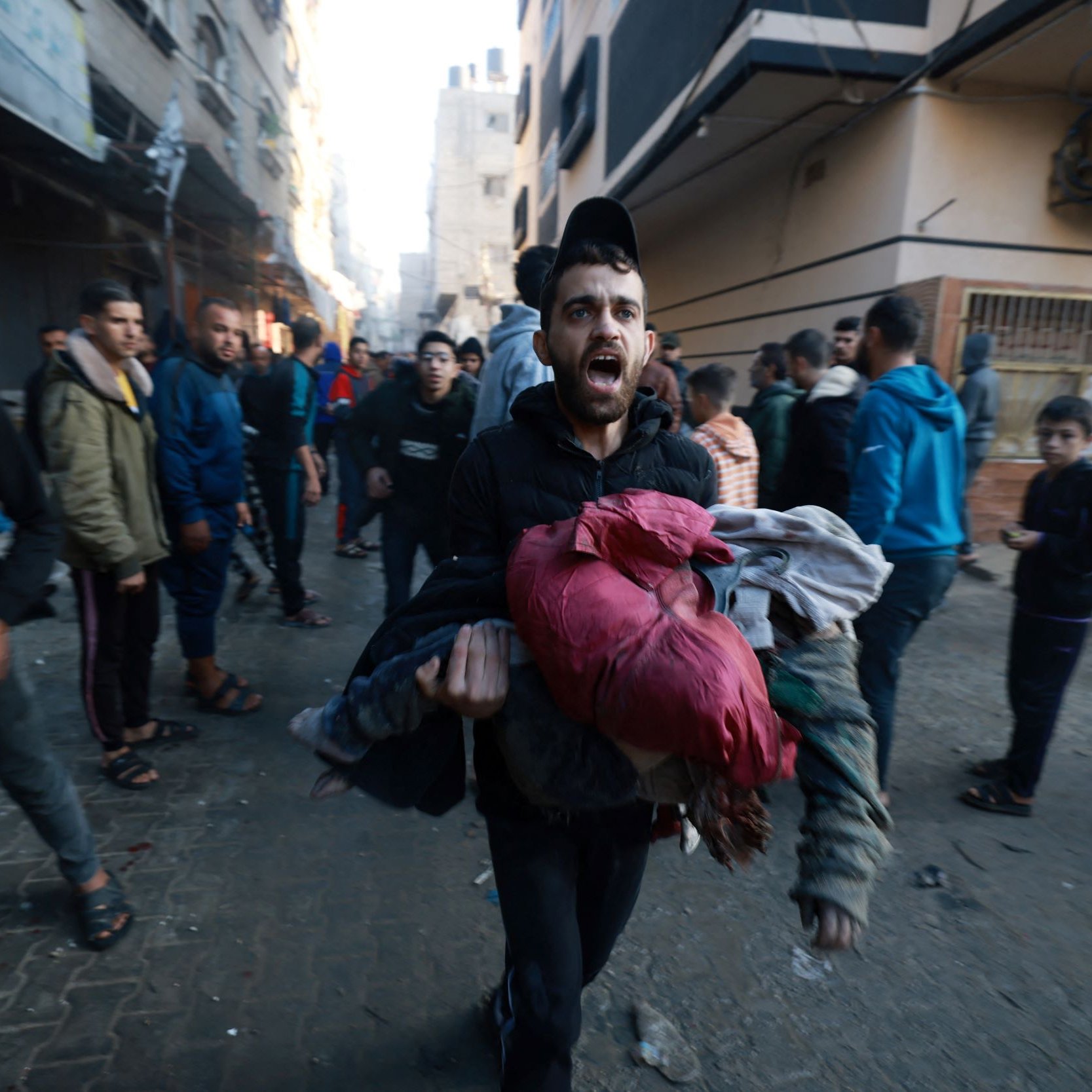 L'image montre une scène urbaine animée où un homme, visiblement en détresse, transporte des vêtements ou des objets dans ses bras. Il semble crier ou exprimer une émotion forte, tandis que d'autres personnes marchent autour de lui. L'environnement est urbain, avec des bâtiments en arrière-plan qui semblent endommagés ou en délabrement. L'atmosphère de l'image suggère une situation de crise ou d'urgence, avec une foule qui se déplace rapidement.