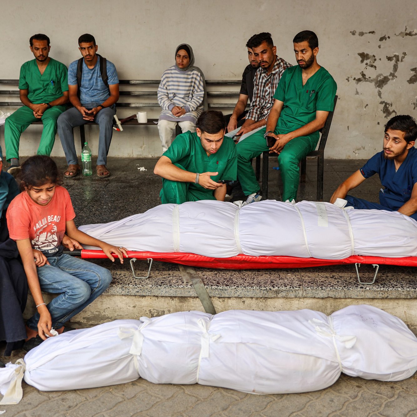 L'immagine mostra un gruppo di persone in uno spazio pubblico. Alcuni individui indossano camici verdi, suggerendo che potrebbero essere operatori sanitari. In primo piano, ci sono due corpi avvolti in lenzuola bianche. Alcuni membri del gruppo sembrano essere in uno stato di lutto, mentre altri osservano la scena in modo serio. L'atmosfera complessiva è di tristezza e riflessione.