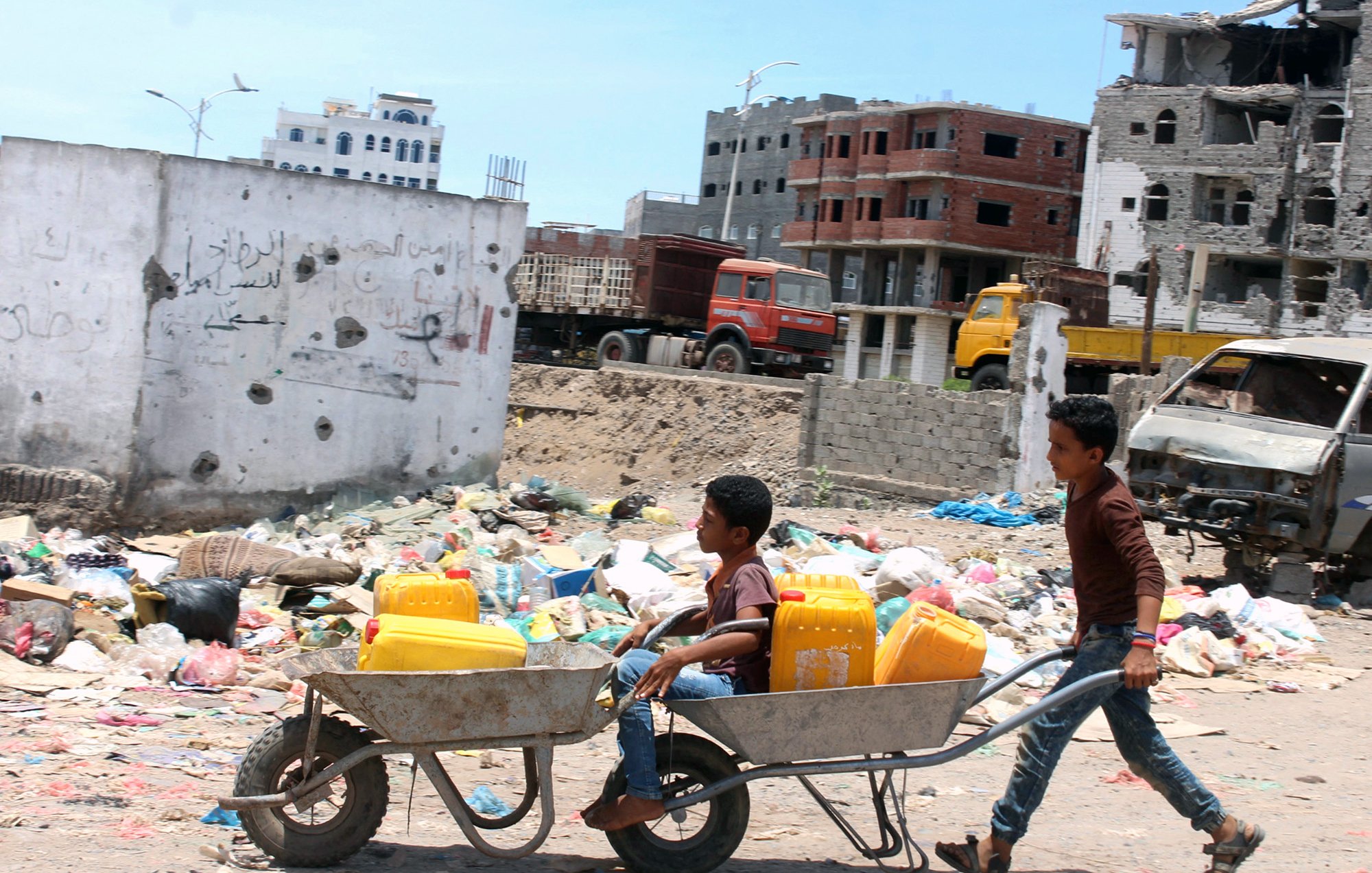تظهر في الصورة مشهدًا مأساويًا يعكس واقعًا صعبًا. يوجد طفلان في منطقة تملأها النفايات. أحد الأطفال يدفع عربة تحتوي على دلوين، بينما يجلس الطفل الآخر في العربة. خلفهم، يمكن رؤية مباني مدمرة وأخرى قيد الإنشاء، مما يعكس تأثير النزاع أو الكوارث على هذه المنطقة. الأجواء تبدو مشمسة، لكن المشهد العام يوحي بالفقر والمعاناة.