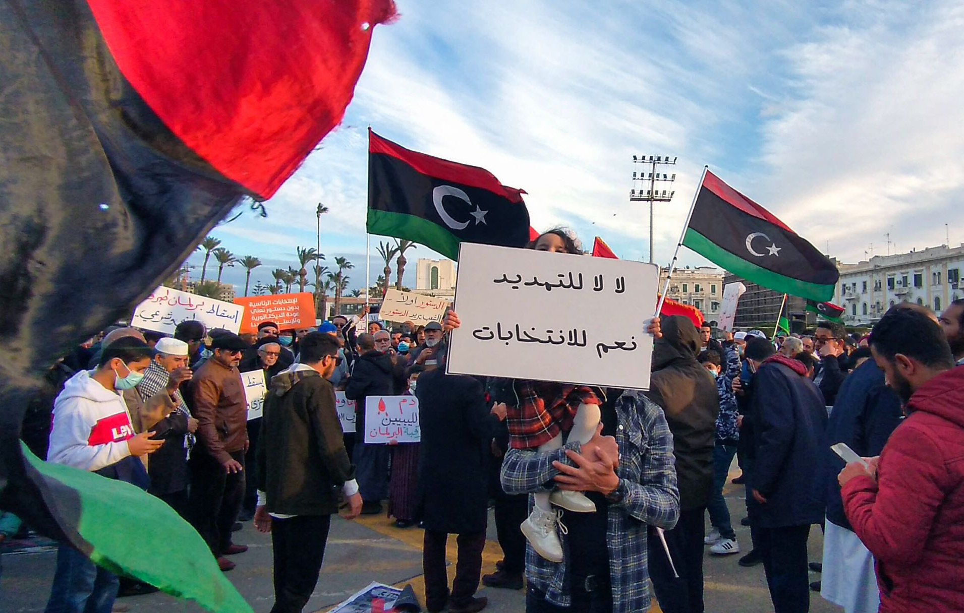 تظهر الصورة مشهداً من تظاهرة في مكان عام، حيث يحتشد الناس حاملين لافتات تتضمن عبارات تتعلق بالحرية والانتخابات. في الخلفية، ترفرف الأعلام الليبية بألوانها الأسود والأحمر والأخضر، مما يدل على الهوية الوطنية. يبدو أن الحشد يتنوع بين الرجال والنساء، وبعضهم يرتدي الكمامات، مما يشير إلى الحرص على الصحة العامة. السماء في الخلفية تظهر مع تزاحم الغيوم، مما يضيف جواً مميزاً إلى البيئة المحيطة بالتجمع.