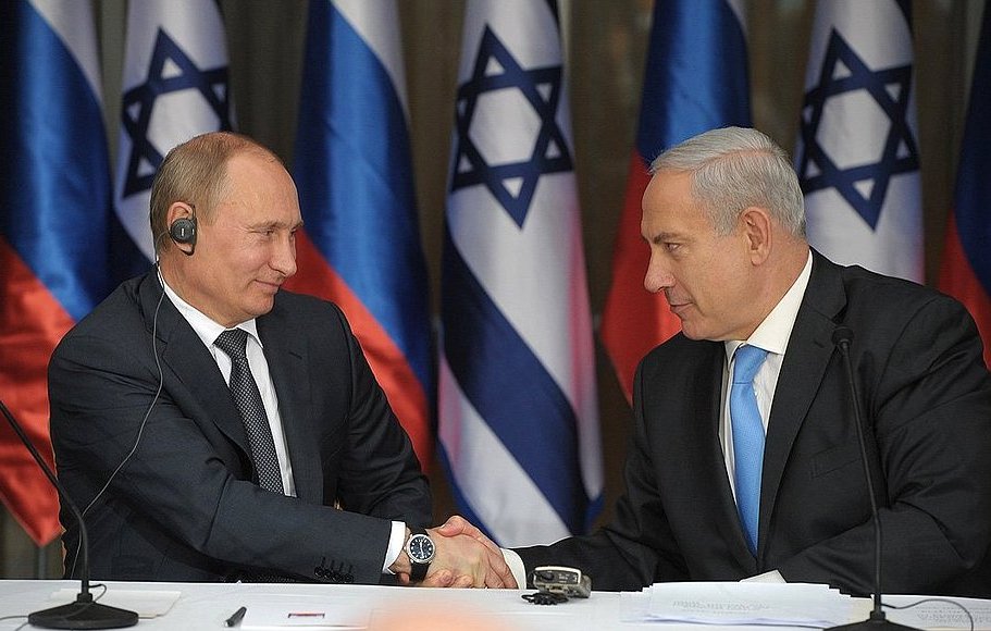 L'image montre deux hommes en costume, se serrant la main, dans un cadre formel. À leur arrière-plan, on peut voir des drapeaux de la Russie et d'Israël. L'un des hommes, à gauche, semble être en train de porter un écouteur, tandis que l'autre, à droite, regarde attentivement. L'ambiance suggère un moment de coopération ou d'accord.