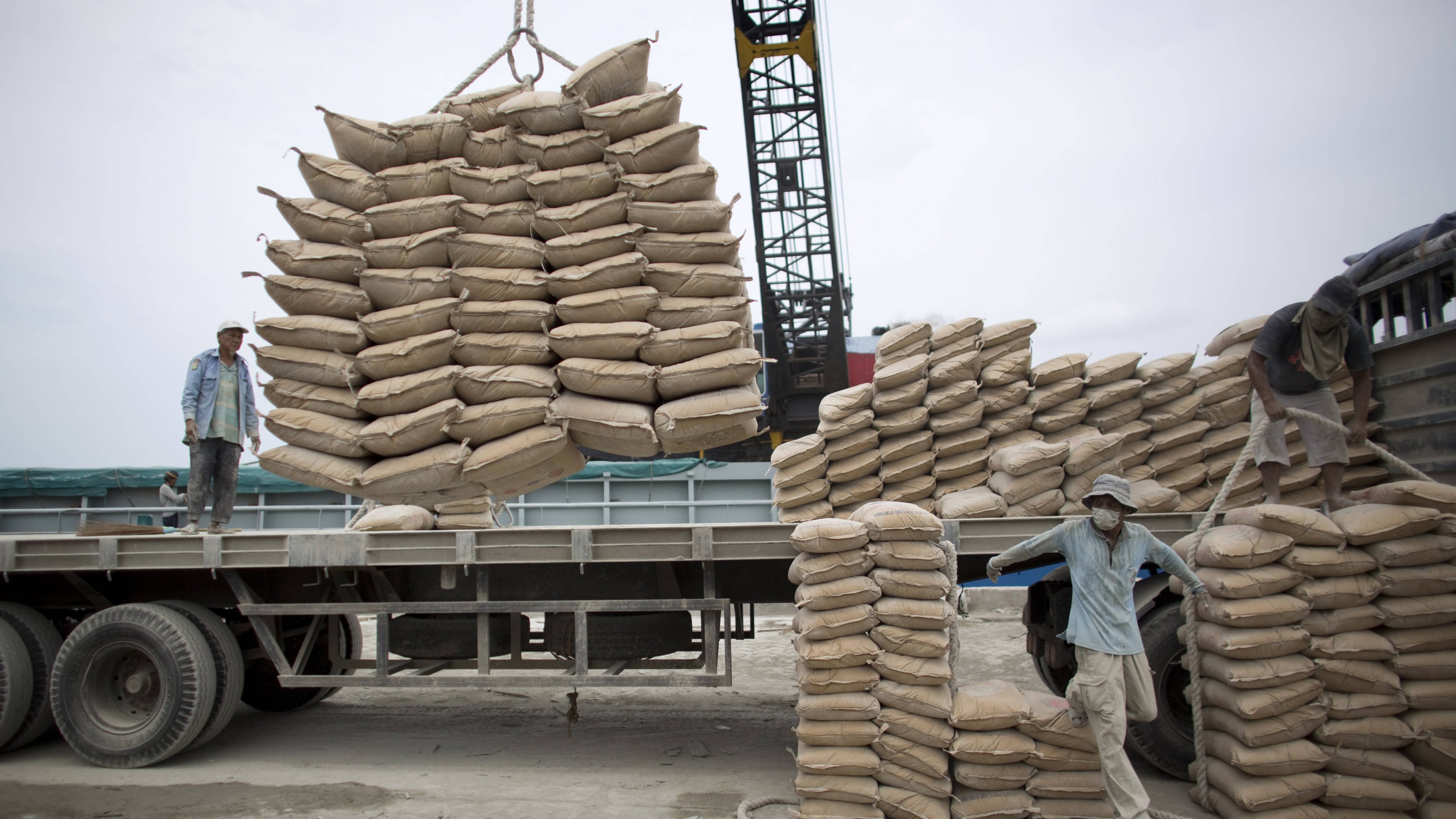 تظهر الصورة عمالًا يقومون بتحميل أكياس من مواد معينة على شاحنة. يبدو أن الأكياس مكدسة بعناية على الشاحنة، حيث يتم رفع بعضها باستخدام رافعة. يتركز العمل على تنظيم الأكياس وضمان تحميلها بشكل صحيح لتسهيل النقل. السماء تبدو غائمة، مما يوحي بأن العمل يتم في يوم غير مشمس.