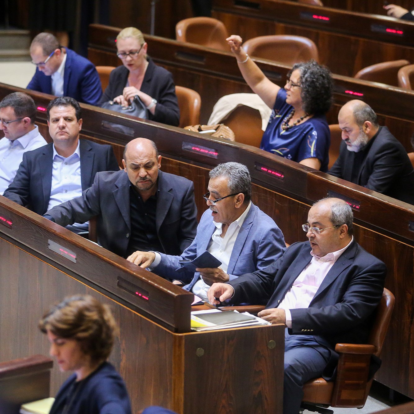 L'image montre une scène à l'intérieur d'une assemblée, probablement dans un parlement. On peut voir plusieurs personnes assises dans des rangées, en train de discuter ou de prendre des notes. Certaines semblent attentives, tandis que d'autres sont engagées dans des conversations. L'environnement est formel, avec des sièges en bois et un éclairage tamisé. Cette composition indique un moment de débat ou de discussion politique.