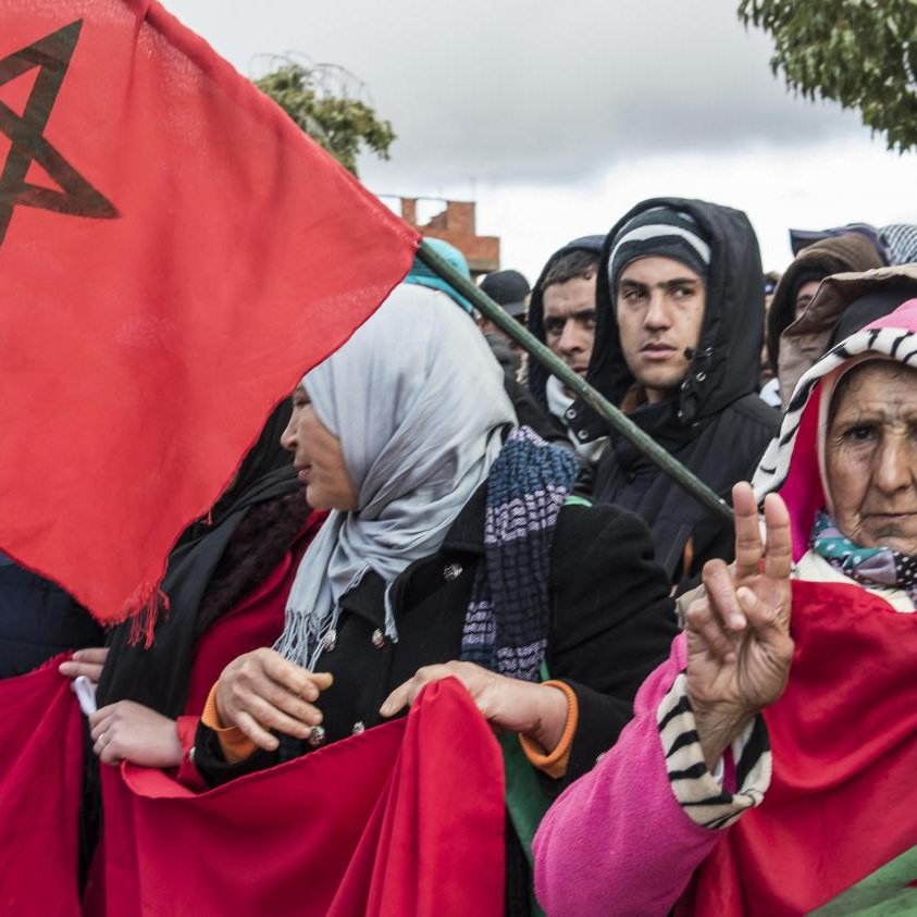 L'image montre une foule rassemblée, avec des gens portant des drapeaux marocains. Parmi eux, on peut voir une femme âgée qui fait un signe de paix avec sa main. Les participants semblent engagés et soutiennent une cause commune, probablement en lien avec leur identité ou des revendications sociales. Leur expression et leur posture suggèrent une forte détermination et un esprit de solidarité. Il y a une atmosphère de mobilisation collective.