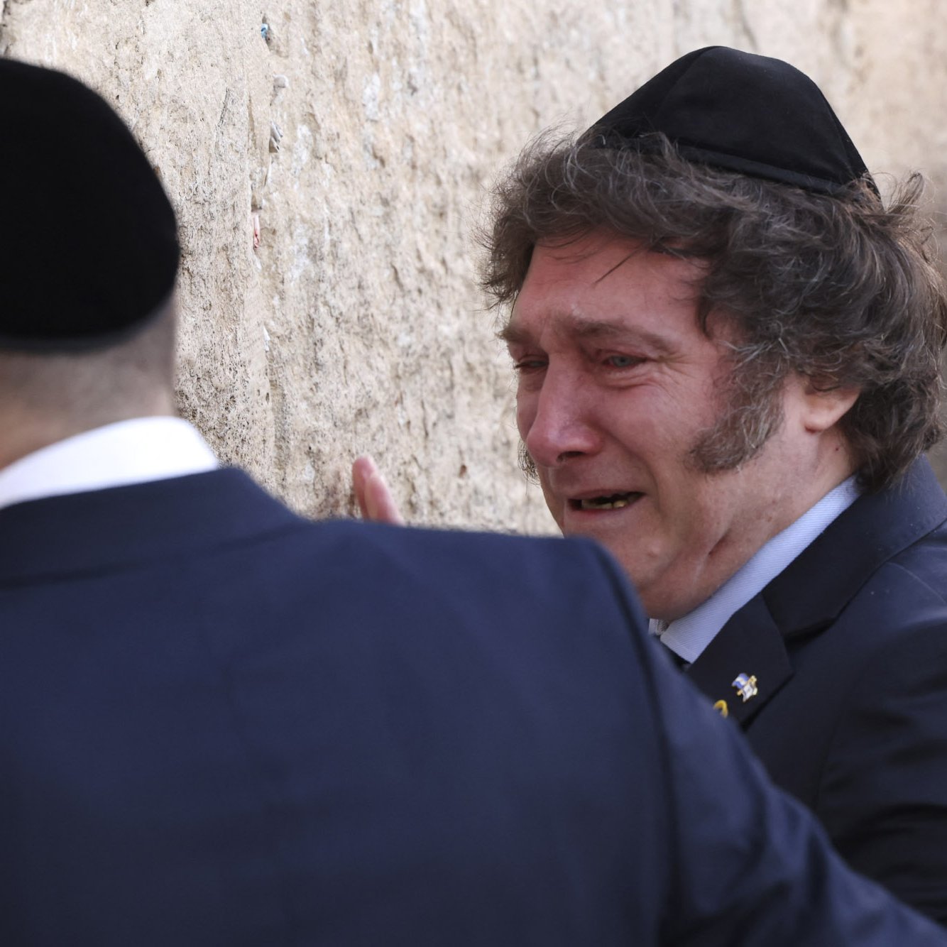 L'image montre deux hommes en train de prier près d'un mur. L'un d'eux, vêtu d'un costume sombre avec une kippa sur la tête, semble pleurer, tandis que l'autre homme, en habit traditionnel, se tient près de lui, peut-être en train de lui offrir du soutien. Le mur derrière eux est de pierre, et l'atmosphère paraît émotive et solennelle.