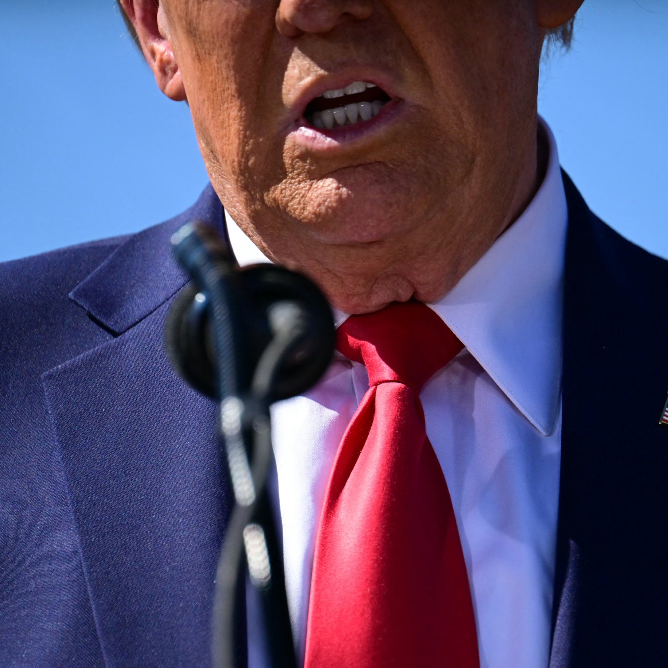 تظهر في الصورة جزء من شخص يرتدي بدلة زرقاء وقميص أبيض مع ربطة عنق حمراء. وقفت الكاميرا بالقرب من الشخص مما يبرز الفم والرقبة، ويظهر أيضًا الميكروفون الذي يتحدث من خلاله. الخلفية زرقاء، مما يشير إلى أن الحدث قد يتم في مكان خارجي.