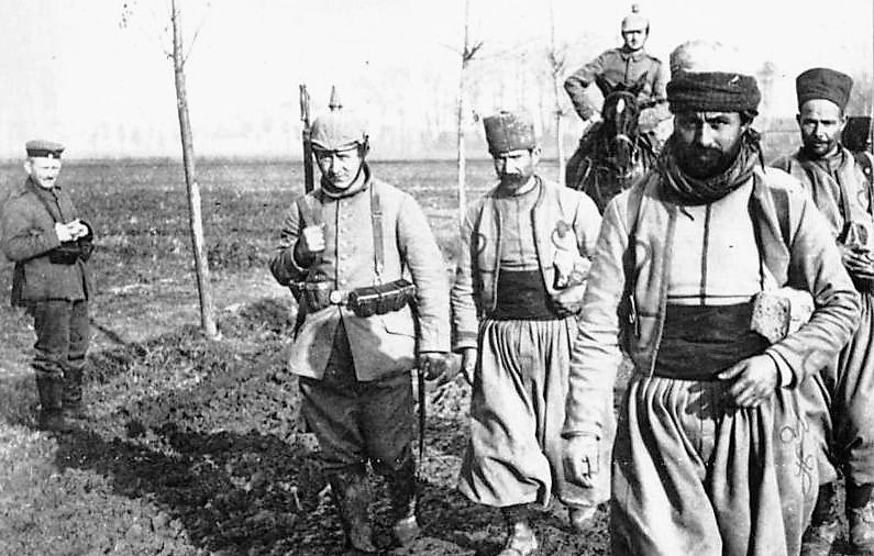 در این تصویر، گروهی از سربازان در حال راه رفتن در یک منطقه باز و مملو از گل و لای هستند. چند نفر از آن‌ها یونیفورم‌هایی به تن دارند که شامل شلوارهای گشاد و کلاه‌های خاص است. یکی از سربازان در حال حمل سلاح است و دیگری دستش را در جیبش قرار داده است. در پس‌زمینه، درختانی دیده می‌شوند که به تازگی کاشته شده‌اند. همچنین، یک نفر دیگر در گوشه تصویر ایستاده و به سمت گروه نگاه می‌کند. این تصویر به نظر می‌رسد که از یک دوره تاریخی خاص گرفته شده باشد و وضعیت نظامی را به نمایش می‌گذارد.