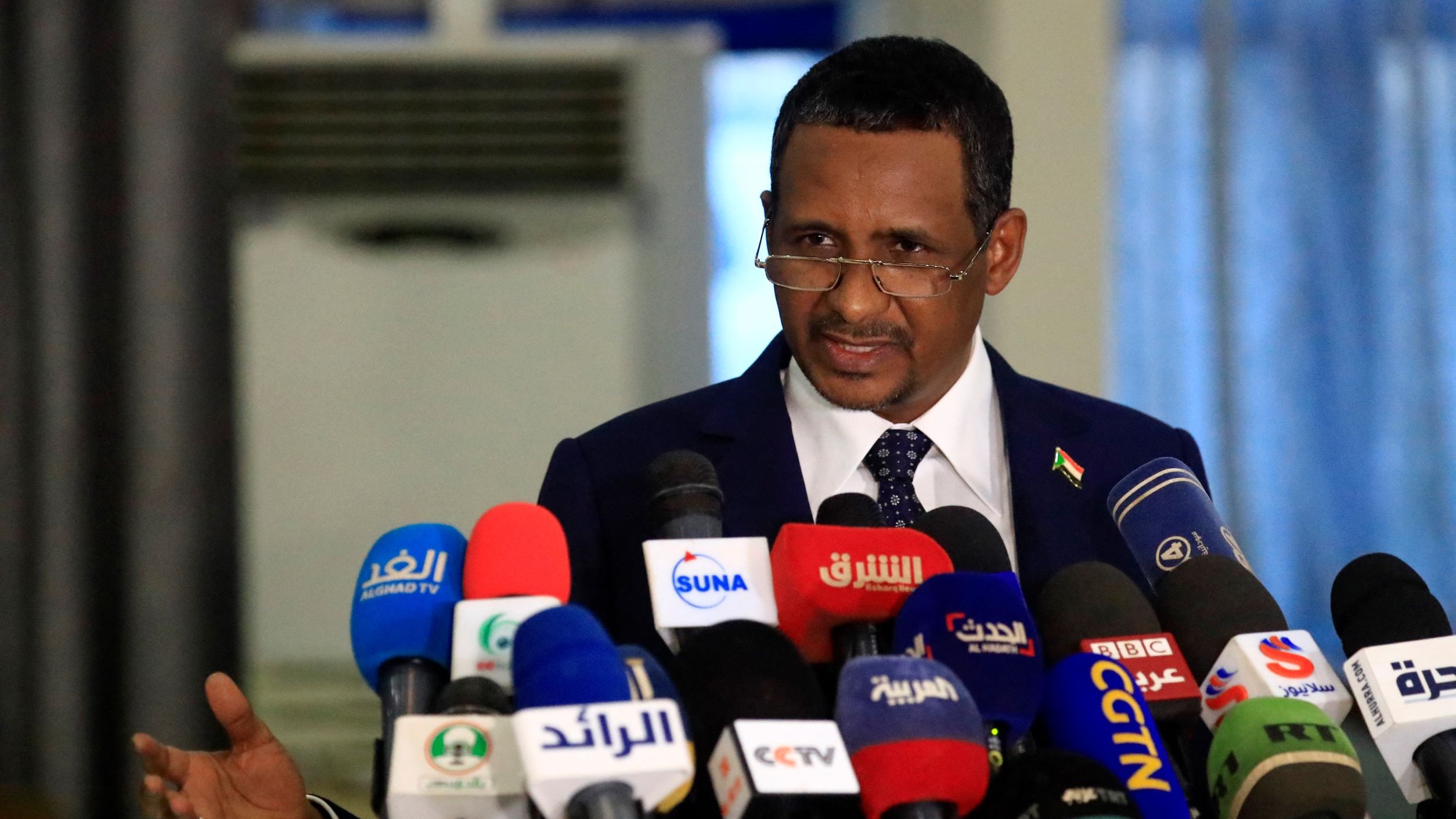 L'image montre un homme en costume, se tenant devant un grand regroupement de microphones provenant de différentes chaînes de télévision et médias. Il semble être en train de faire une déclaration ou un discours, avec des gestes expressifs. L'environnement est formel, suggérant une conférence de presse ou un événement politique.
