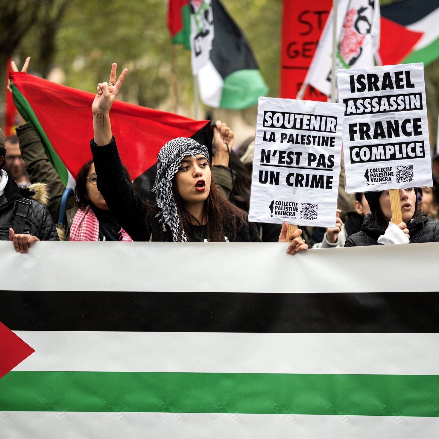 تظهر الصورة مجموعة من المتظاهرين في شارع، يحملون لافتات وشعارات تتعلق بفلسطين. يوجد علم فلسطيني في مقدمة المظاهرة، ويتوسط الحشد شخص يرفع ذراعيه بعلامة النصر. اللافتات تحمل عبارات تدعو لدعم فلسطين وتندد بفعل الإسراء وتتهم فرنسا بالتواطؤ. المشهد يعكس شعورًا قويًا بالتضامن مع القضية الفلسطينية.