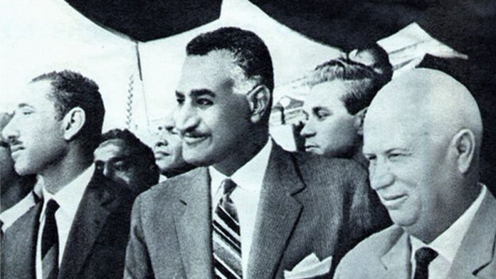 الصورة تظهر ثلاثة رجال يقفون في حدث رسمي. الرجل في المنتصف يرتدي بدلة أنيقة مع ربطة عنق، ويظهر عليه مظهرٌ واثق. الشخصان الآخران على جانبيه يبدو أنهم يرتدون ملابس رسمية أيضًا. يبدو أن البيئة من حولهم تشير إلى مناسبة مهمة. للحظة، يبدو أنهم يتحدثون أو يتبادلون الآراء حول موضوع ما. الصورة تحمل طابعًا تاريخيًا، ربما تشير إلى فترة معينة في السياسة أو العلاقات الدولية.