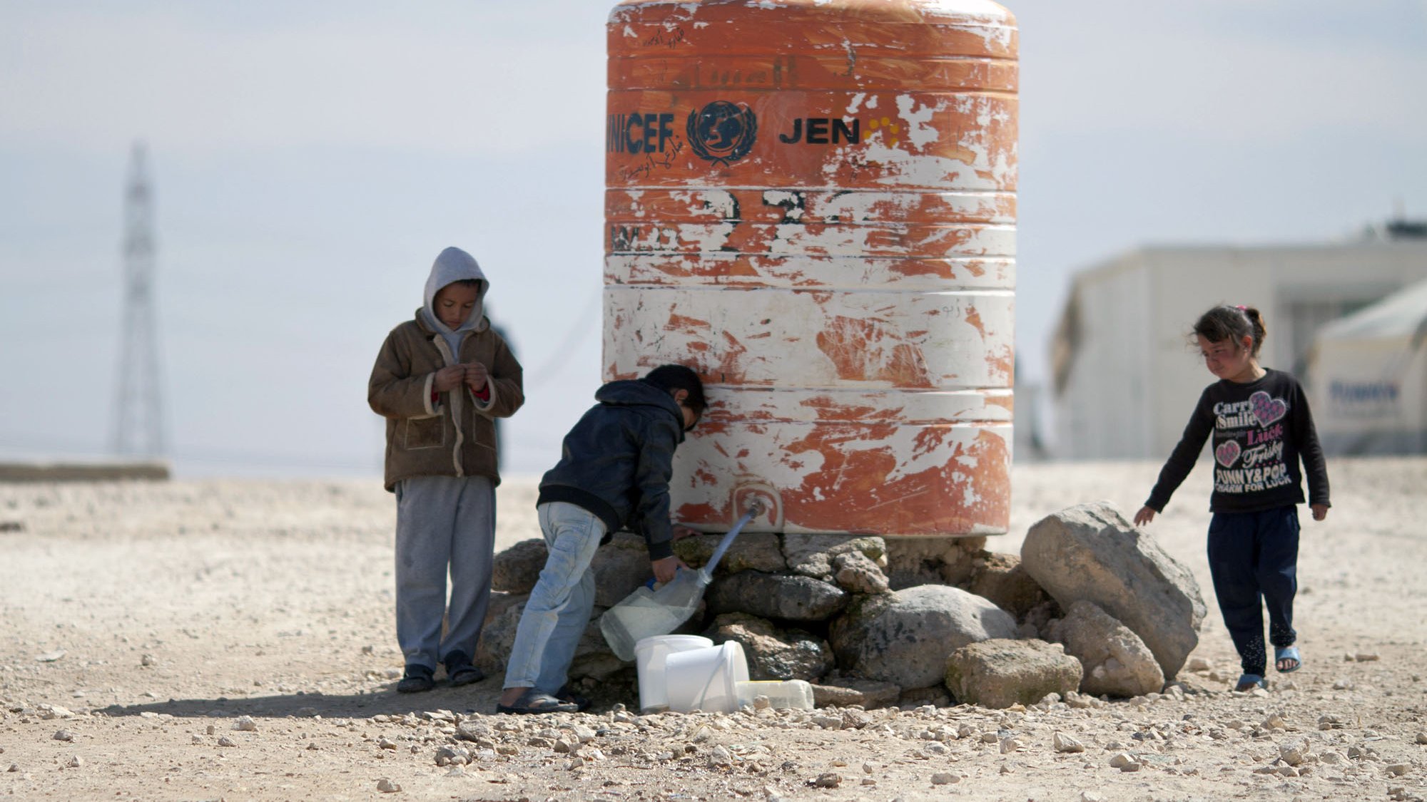 تظهر الصورة مجموعة من الأطفال بالقرب من خزان ماء كبير مُسجّل عليه علامة الأمم المتحدة. يقف اثنان من الأطفال بجانب الخزان، حيث يبدو أحدهم مشغولاً في فحص شيء ما، بينما يقوم الآخر بسكب الماء في وعاء. هناك طفل ثالث يلعب بجانبهم. الخلفية تبدو قاحلة مع بعض الصخور والأرض الرملية، مما يشير إلى بيئة جافة.