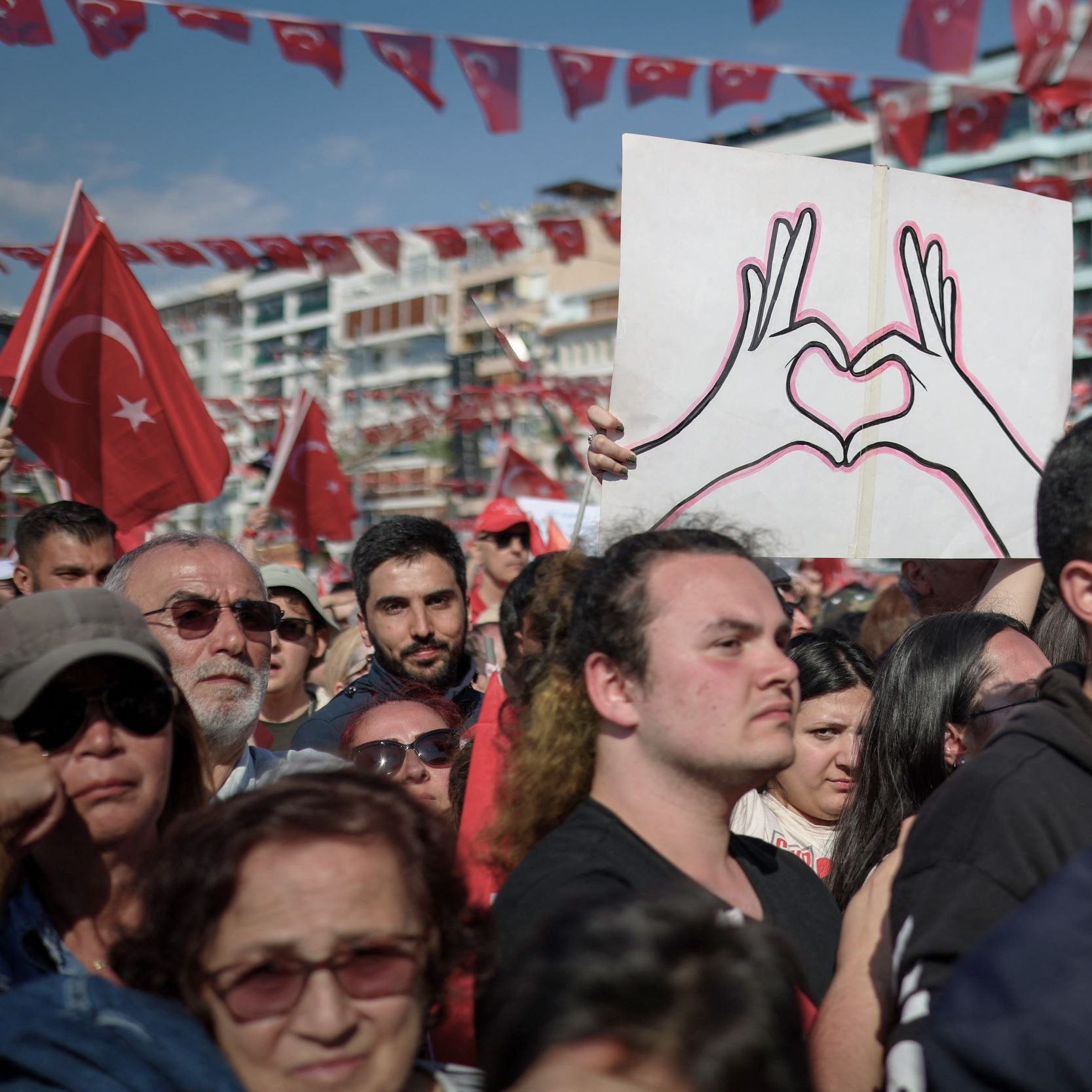 در این تصویر، تجمعی از مردم دیده می‌شود که برخی از آن‌ها پرچم‌های قرمز رنگ ترکیه را در دست دارند. در پس‌زمینه، یک پلاکارد بزرگ با طراحی دو دست که همدیگر را درحال ایجاد شکل قلب نشان می‌دهد، قرار دارد. افراد حاضر در جمعیت به نظر می‌رسد که توجهشان به پلاکارد جلب شده و فضای کلی تجمع نشان‌دهنده شور و شوق آن‌ها است. تعدادی از مردم با چهره‌های مختلف در تصویر وجود دارند، که نشان‌دهنده تنوع شرکت‌کنندگان در این مراسم است.