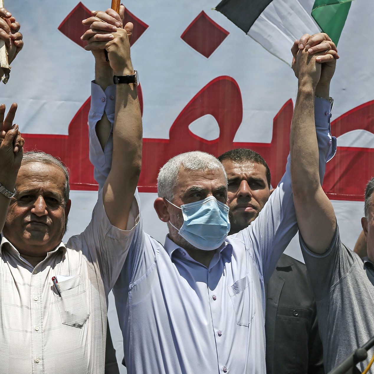 The image depicts a group of men at a public gathering or protest. They are raising their hands in the air, with some holding sticks or flags. The man at the center is wearing a mask and appears to be leading or participating actively in the event. In the background, there is a large banner in Arabic, suggesting a political or social message. The mood of the event seems to be one of solidarity or protest, with participants visibly engaged.