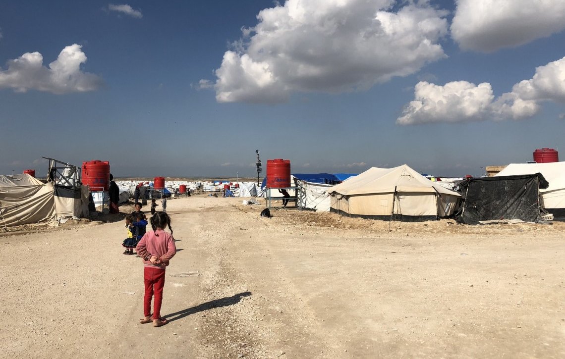 L'image montre un camp de réfugiés avec des tentes disposées de part et d'autre d'un chemin de terre. Au fond, on aperçoit des nuages blancs dans un ciel bleu. Des enfants sont présents, certains semblent jouer ou se déplacer dans le camp. Les abris sont principalement en toile, avec quelques réservoirs d'eau visibles. L'atmosphère paraît calme malgré les conditions de vie apparentes de ces lieux.