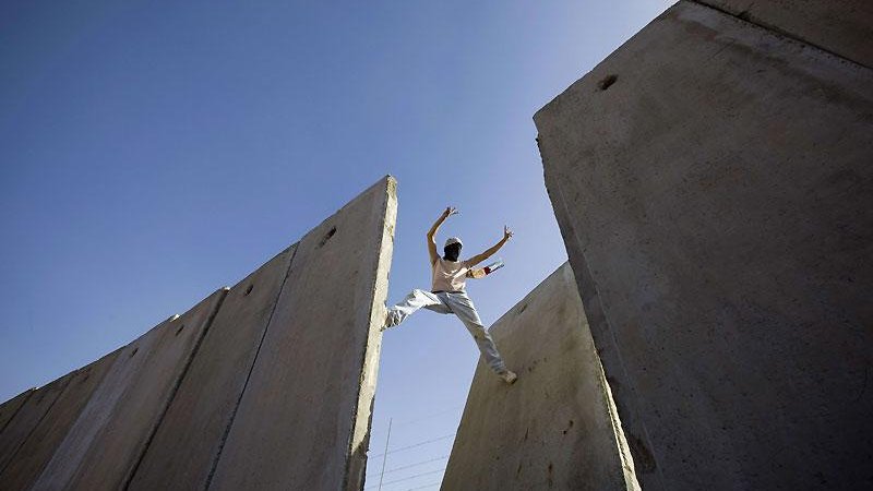 L'image montre une personne qui effectue une sorte d'acrobatie ou de saut entre deux grandes structures en béton. Le ciel est bleu, ce qui donne une impression d'ampleur et de liberté. La personne semble en mouvement, exploitant l'espace entre les murs. Cette scène peut évoquer des thèmes tels que le dépassement des obstacles ou la recherche de la liberté.