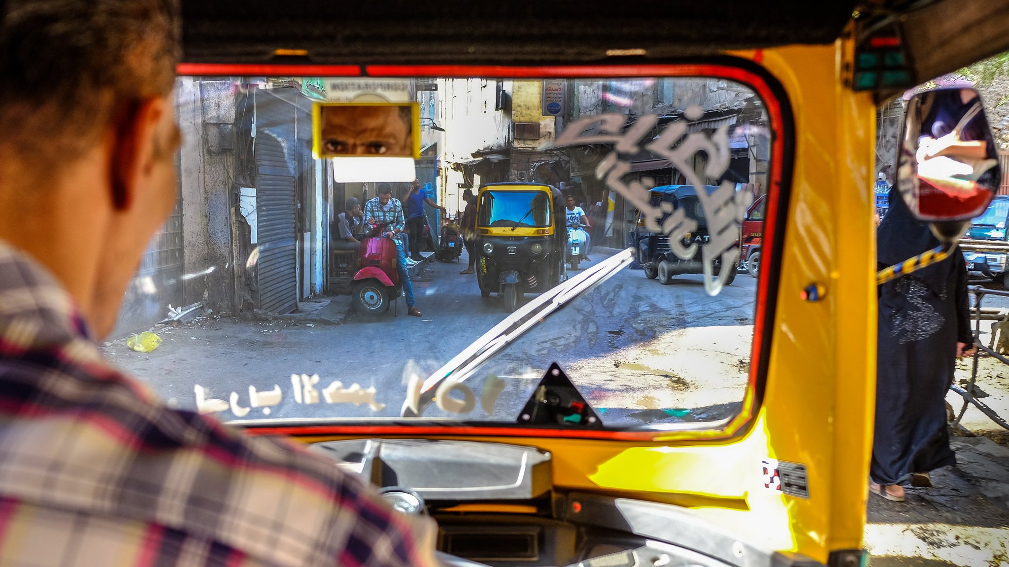 L'image montre la vue depuis l'intérieur d'un tuk-tuk, avec un conducteur visible en premier plan. On aperçoit des rues étroites et animées, avec d'autres véhicules, y compris un autre tuk-tuk. Des bâtiments anciens bordent la rue, et il y a des détails comme des affiches et des éléments de la vie urbaine qui ajoutent du caractère à l'environnement. La lumière naturelle éclaire la scène, créant une atmosphère vivante et dynamique.
