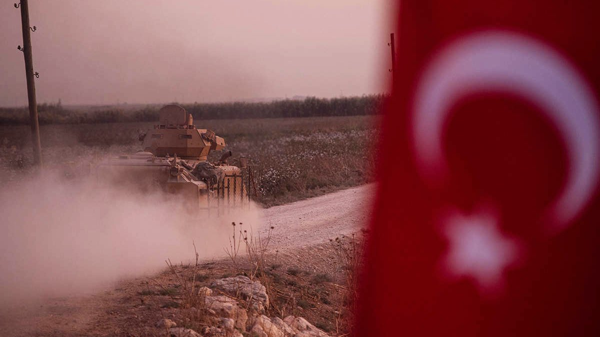 The image depicts a military tank moving along a dirt road, creating a cloud of dust. In the foreground, part of a Turkish flag is visible, featuring a white star and crescent on a red background. The landscape appears to be rural, possibly with fields or open land in the background, and the lighting suggests either dawn or dusk, giving a somewhat dramatic atmosphere to the scene.
