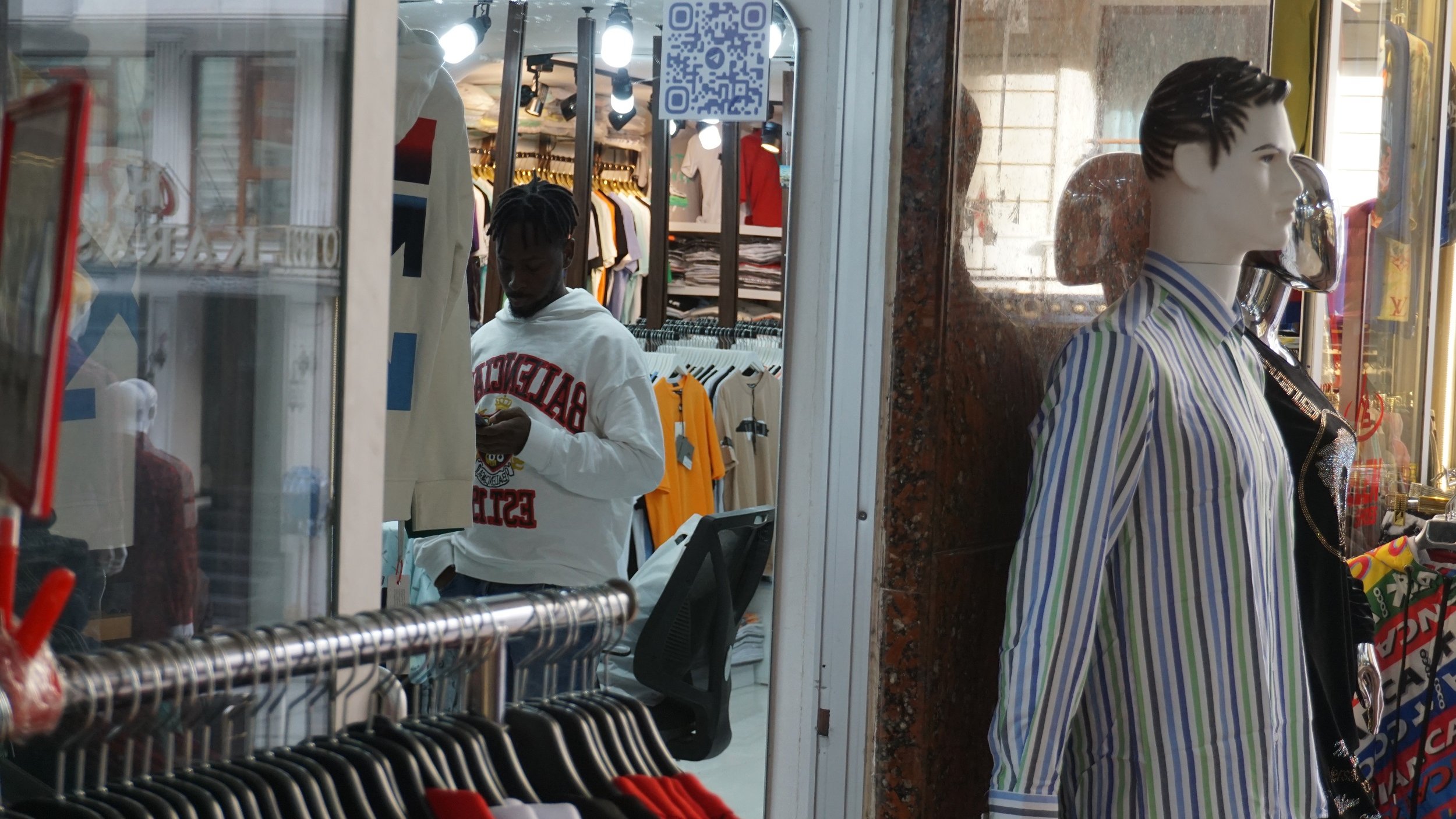 L'image montre un magasin de vêtements avec des rangées de vêtements accrochés, notamment des t-shirts et des chemises multicolores. À gauche, un homme portant un sweat-shirt blanc est en train de regarder son téléphone. À droite, un mannequin habillé d'une chemise à rayures est exposé dans la vitrine. On peut aussi apercevoir un code QR accroché à l'intérieur du magasin. L ambiance est typique d'un espace commercial animé.