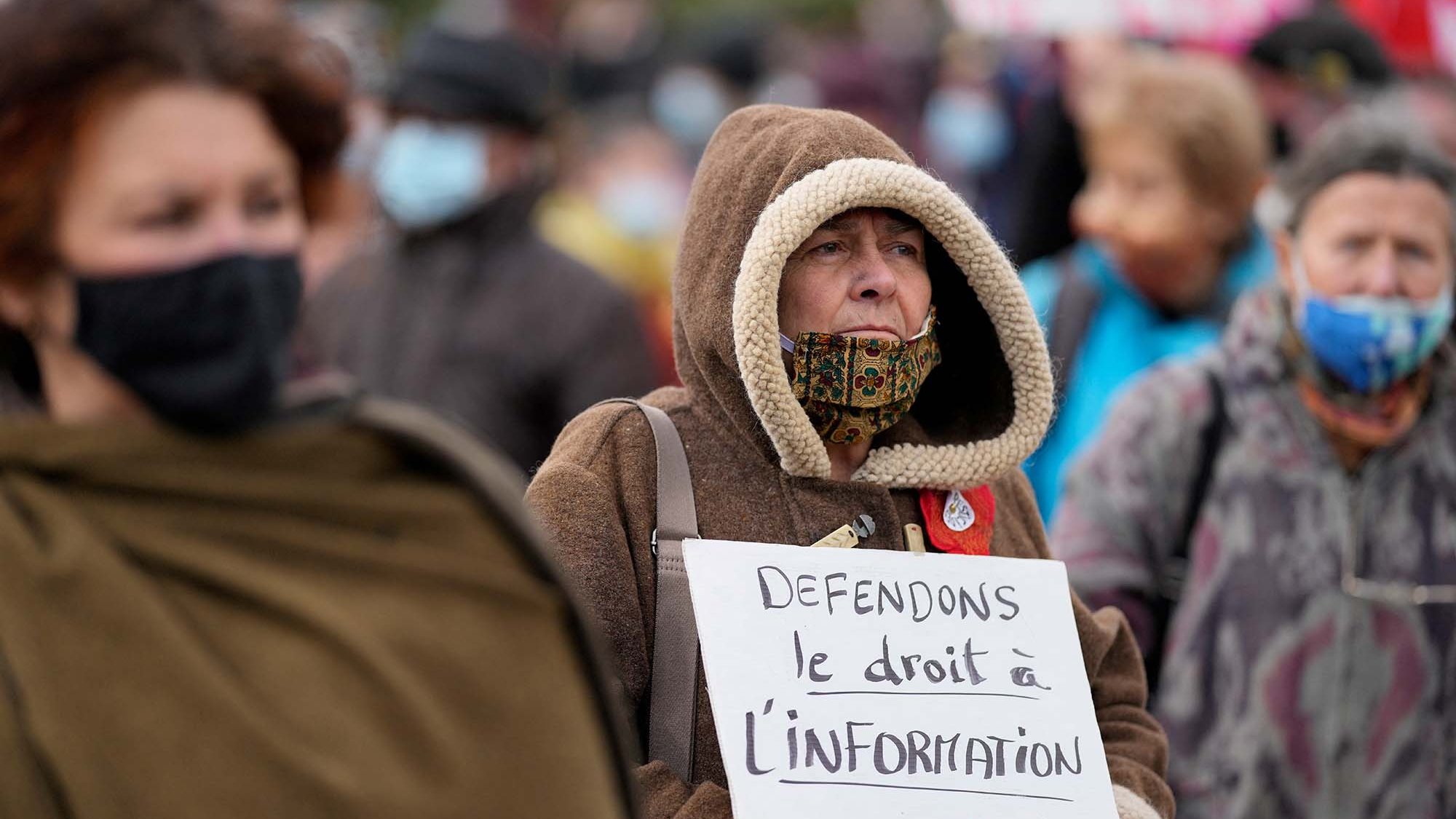 L'image montre un groupe de personnes rassemblées lors d'une manifestation. Au premier plan, une femme âgée porte une grande capuche et un masque, tenant une pancarte sur laquelle il est écrit : "Défendons le droit à l'information". Les autres manifestants sont flous en arrière-plan, mais on peut voir qu'ils portent également des masques. L'atmosphère semble sérieuse et engagée, avec des personnes qui militent pour leurs droits.