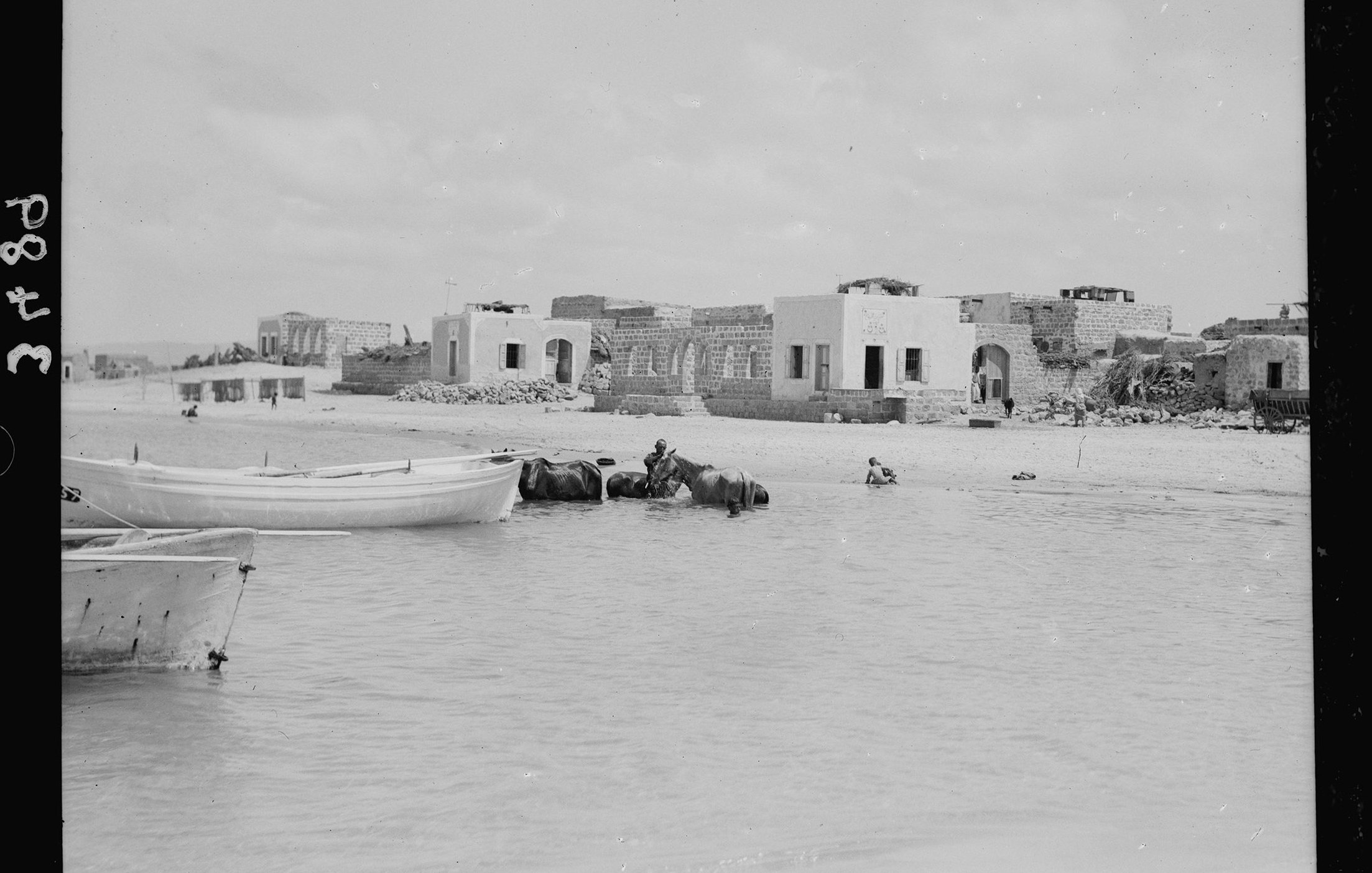 L'image montre une scène maritime où plusieurs bateaux, probablement des barques, sont amarrés au bord d'une eau peu profonde. En arrière-plan, on peut apercevoir des bâtiments en ruines, suggérant une ancienne architecture. À proximité, un homme semble se trouver dans l'eau, tandis qu'un autre est à cheval ou monté sur un animal aquatique. Le ciel est nuageux, apportant une lumière diffuse à l'ensemble de la scène. L'atmosphère semble tranquille et un peu nostalgique, évoquant un passé révolu.