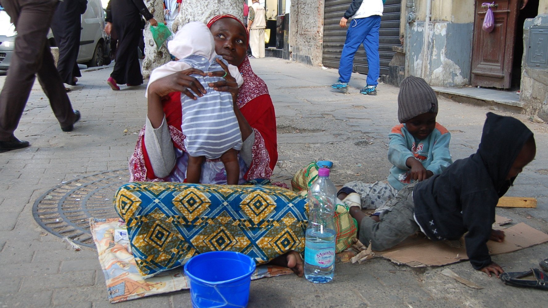The image depicts a scene on a city street where a woman is seated on the ground with a baby in her arms. She appears to be wearing traditional clothing, characterized by vibrant colors and patterns. Nearby, two young children are playing on the pavement; one is sitting in a casual posture, while the other is occupied with an object. There is a blue bucket beside them and a plastic bottle of water. The background includes other people walking by and buildings that suggest an urban environment.