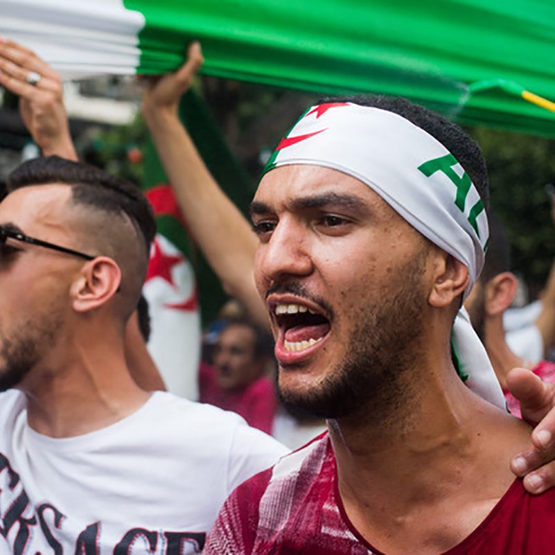 L'image montre des manifestants lors d'une protestation, probablement en lien avec une cause nationale. Deux hommes sont au premier plan : l'un porte un bandana avec des motifs évoquant l'Algérie et semble exprimer de la passion en criant. L'autre homme, à sa gauche, affiche une attitude engagée tout en portant un t-shirt à motifs. En arrière-plan, on peut voir un groupe de personnes brandissant des drapeaux, créant une ambiance de solidarité et de revendication. Les visages sont chargés d'émotion et l'atmosphère paraît énergique.