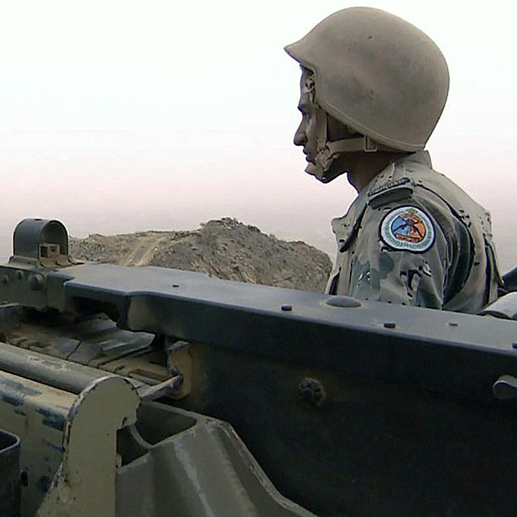 The image depicts a soldier sitting beside a mounted machine gun, which is positioned for observation or potential engagement. The soldier is wearing a helmet and a military uniform adorned with insignia. The background features a foggy or hazy landscape, suggesting a possibly mountainous or rugged terrain. The overall atmosphere conveys a sense of vigilance and readiness in a military setting.
