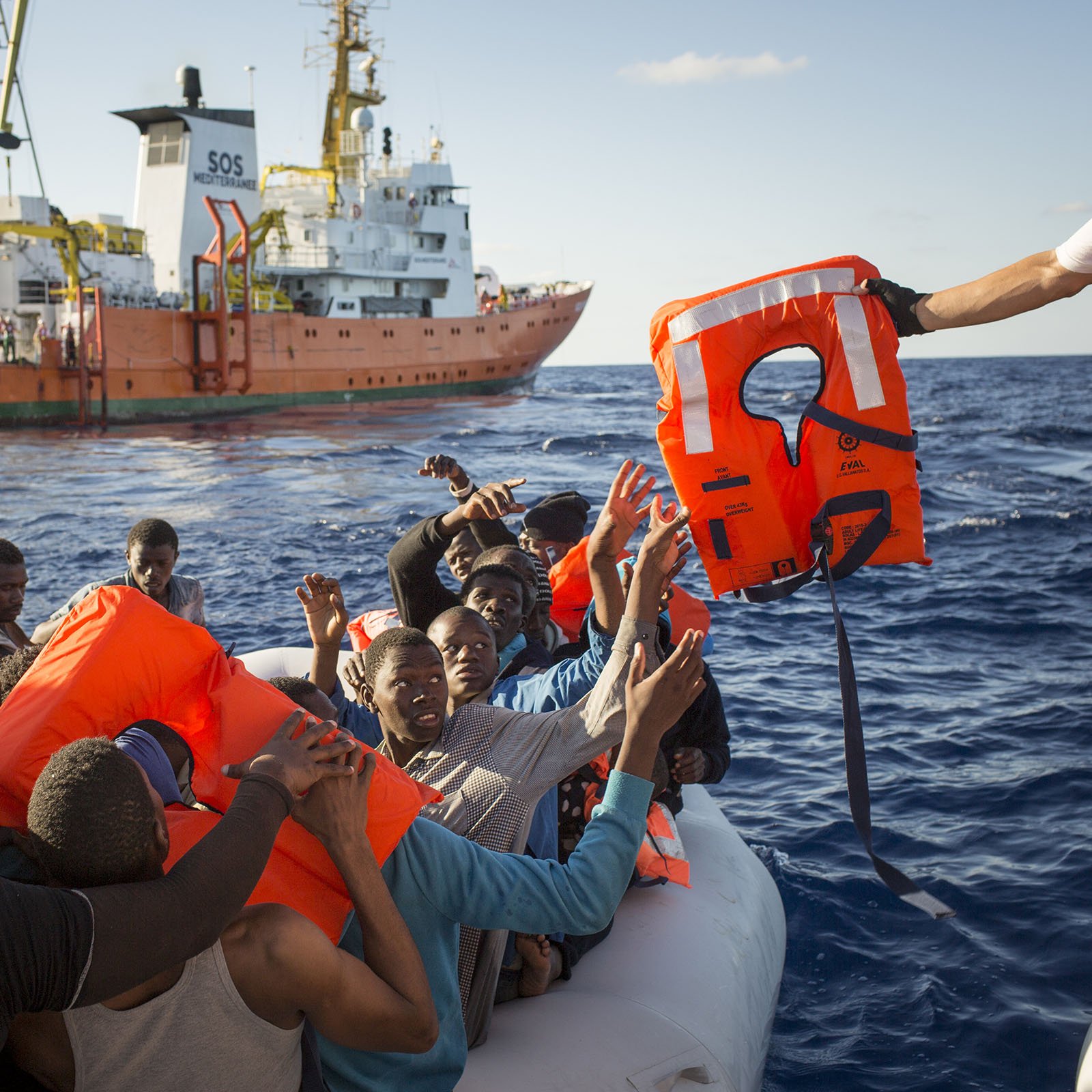 L'image montre une scène en mer où un groupe de personnes se trouve sur un canot pneumatique. Elles semblent en détresse et soulèvent les bras en direction d'un sauveteur qui leur tend une bouée de sauvetage. En arrière-plan, un grand navire de secours est visible, avec des couleurs orange et blanc. L'atmosphère semble tendue, et on peut ressentir l'urgence de la situation. Le ciel est dégagé et l'eau est calme, contrastant avec la gravité du moment.