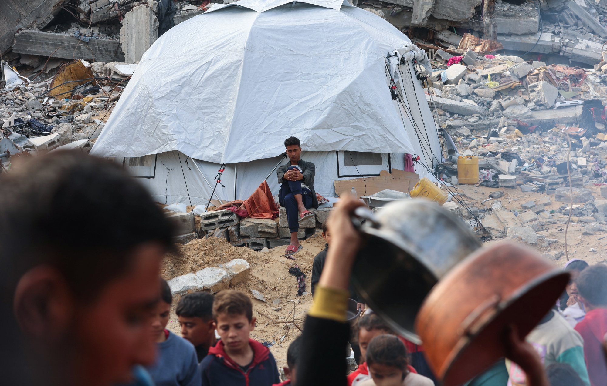 Nell'immagine si vede una tenda bianca in un contesto di distruzione, con rovine e macerie intorno. Un ragazzo è seduto davanti alla tenda, mentre in primo piano ci sono persone che si muovono e tengono stoviglie. L'atmosfera sembra di emergenza e difficoltà.