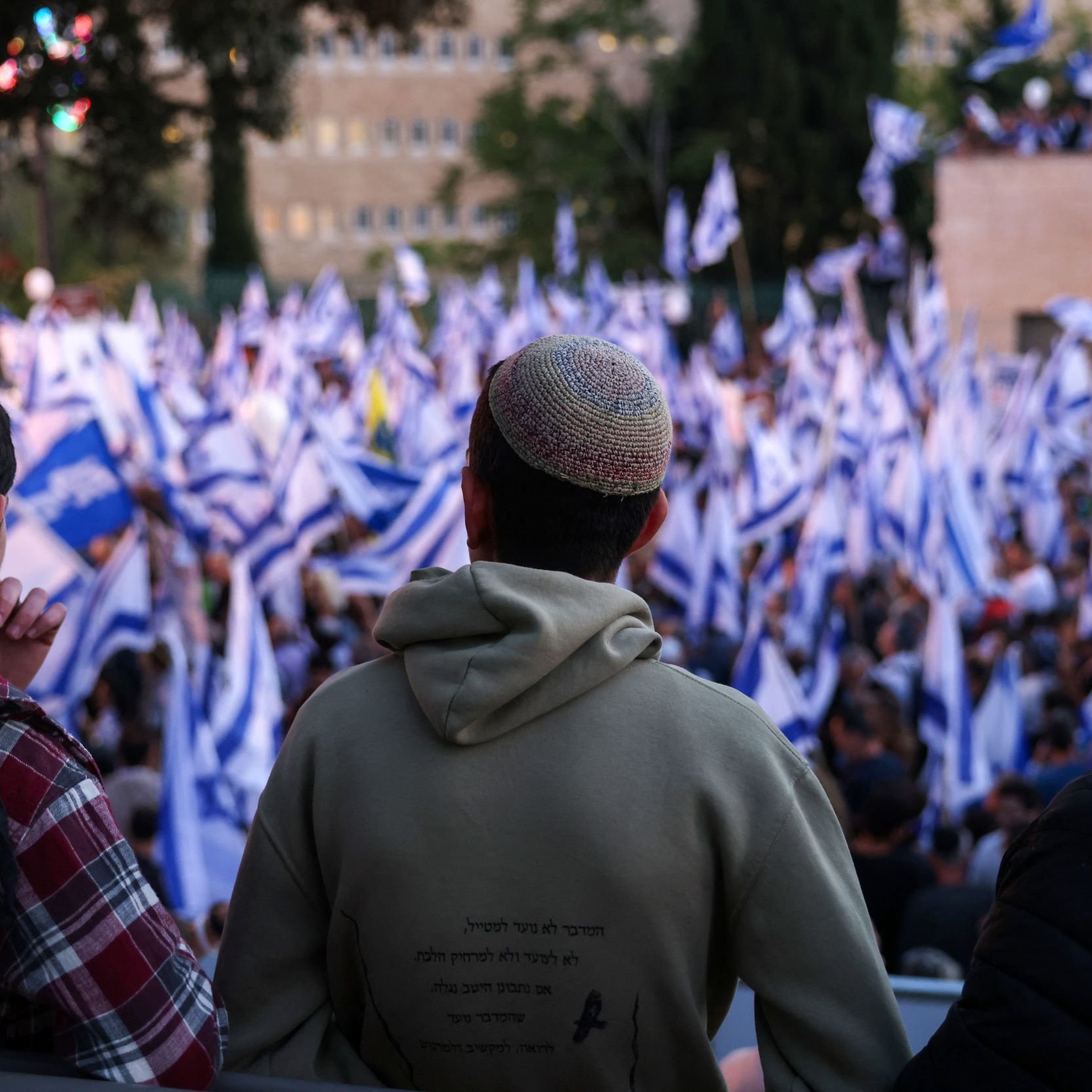 L'image montre un groupe de personnes vu de dos, regardant une manifestation ou un rassemblement. Ils portent des kippas. Au premier plan, on observe un homme en sweat à capuche, tandis que les autres sont légèrement flous. En arrière-plan, une foule dense agite des drapeaux, probablement israéliens, créant une ambiance dynamique. La scène semble se dérouler à l'extérieur, avec des arbres et des bâtiments visibles au loin.