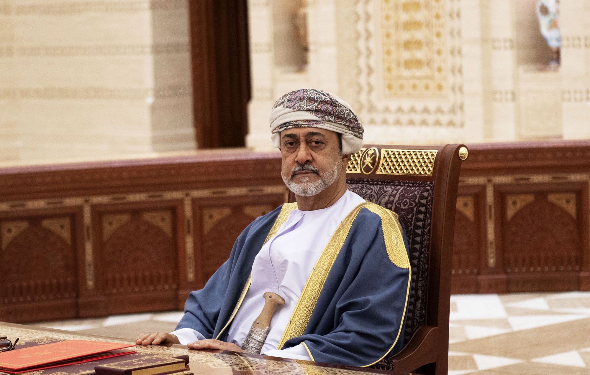 L'image montre un homme assis à un bureau dans un environnement intérieur richement décoré. Il porte un costume traditionnel, comprenant une robe longue et un turban orné. Le décor autour de lui présente des motifs architecturaux détaillés, typiques d'une ambiance élégante. Il semble concentré et sérieux, et le mobilier en bois ajoute à l'atmosphère formelle de la scène.