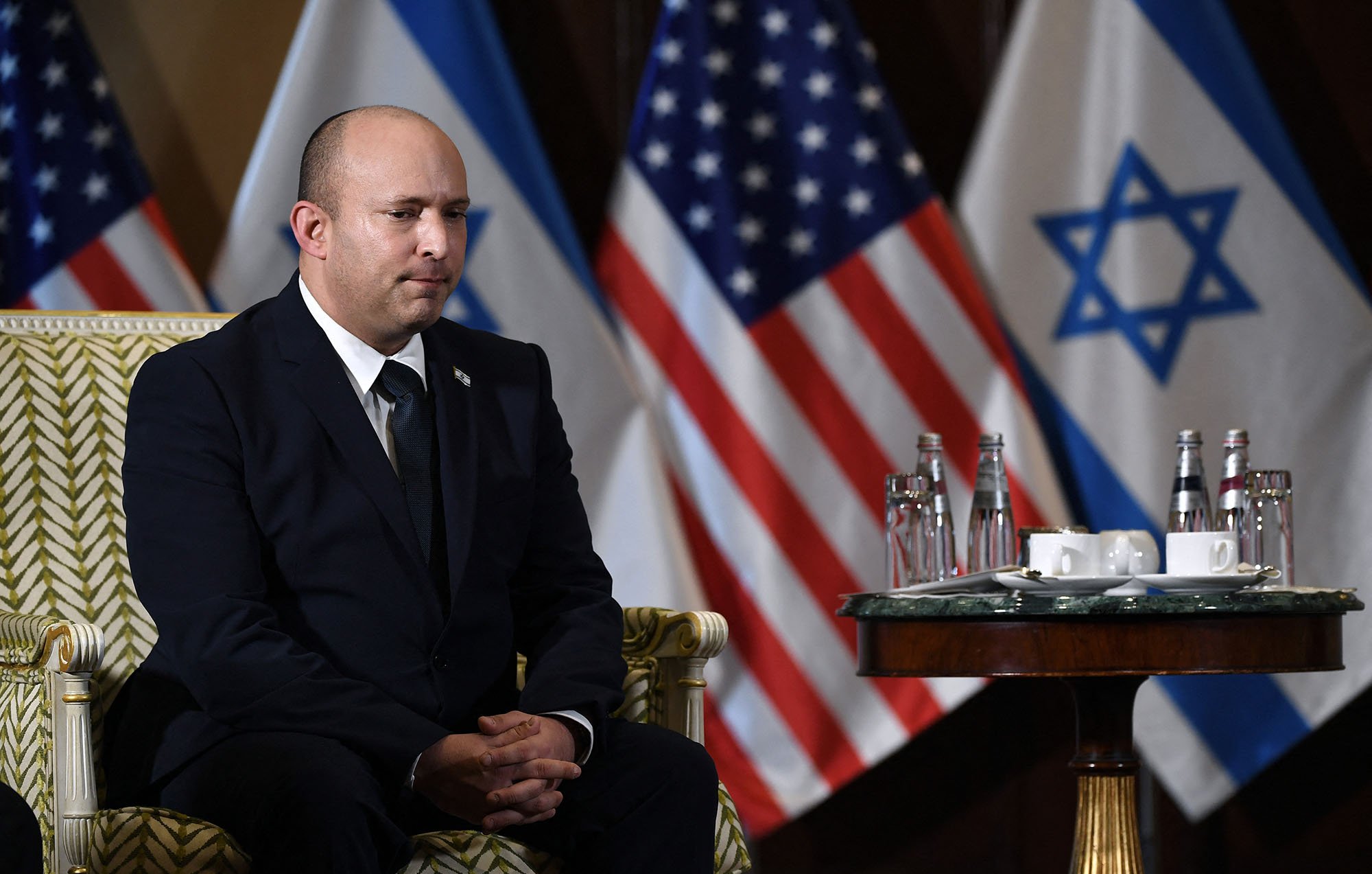 L'image montre un homme assis sur une chaise ornée, vêtu d'un costume sombre. Il a une expression sérieuse et semble concentré. À l'arrière-plan, on peut voir plusieurs drapeaux, dont ceux des États-Unis et d'Israël. Sur la table devant lui, il y a des bouteilles d'eau et des tasses. L'ambiance semble officielle et solennelle.