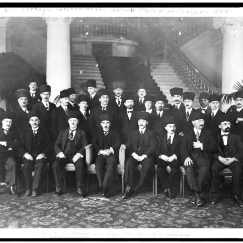L'image présente un groupe d'hommes, probablement d'importance politique ou officielle, assis et debout dans un cadre formel. Ils portent des costumes sombres et des chapeaux traditionnels, et certains ont des moustaches. La scène se déroule dans un intérieur élégant, avec des colonnes décoratives et des plantes en pot en arrière-plan. L'atmosphère dégage une impression de solennité et de cérémonie, suggérant un événement ou une réunion importante.