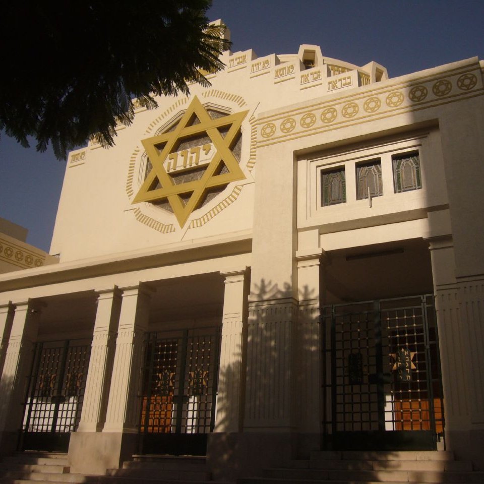 L'image montre un bâtiment architectural avec des éléments néo-classiques. La façade est ornée d'une étoile de David, symbolisant une influence juive. Les fenêtres sont encadrées de détails géométriques et il y a une entrée principale bien marquée. Le bâtiment est éclairé par la lumière du jour, ce qui met en valeur ses décorations. Des arbres sont visibles au premier plan, ajoutant une touche de verdure à la scène.