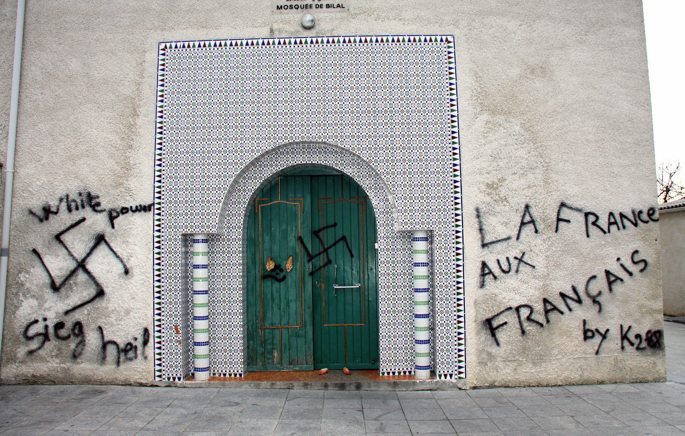 L'image montre un mur extérieur d'une mosquée, orné de carreaux colorés. Sur le mur, il y a des graffitis qui incluent un symbole controversé, représentant une swastika, ainsi que des phrases telles que "White power" et "La France aux Français." La porte d'entrée est en bois, avec des détails architecturaux typiques. L'ensemble de la scène évoque un message de haine et de division.