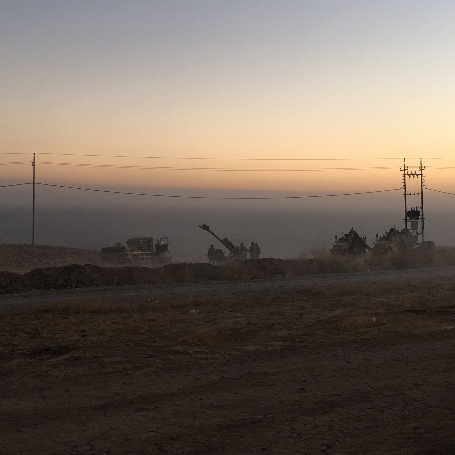 L'image montre un paysage de chantier au crépuscule, avec un ciel teinté de couleurs douces allant du jaune à l'orange. On aperçoit des machines de construction et des travailleurs en silhouettes, entourés de poussière. À l'arrière-plan, on distingue des poteaux électriques. Le sol semble sec et rocailleux, suggérant une zone de travaux en cours. L'atmosphère est calme, mais il y a une impression d'activité et d'effort en cours.