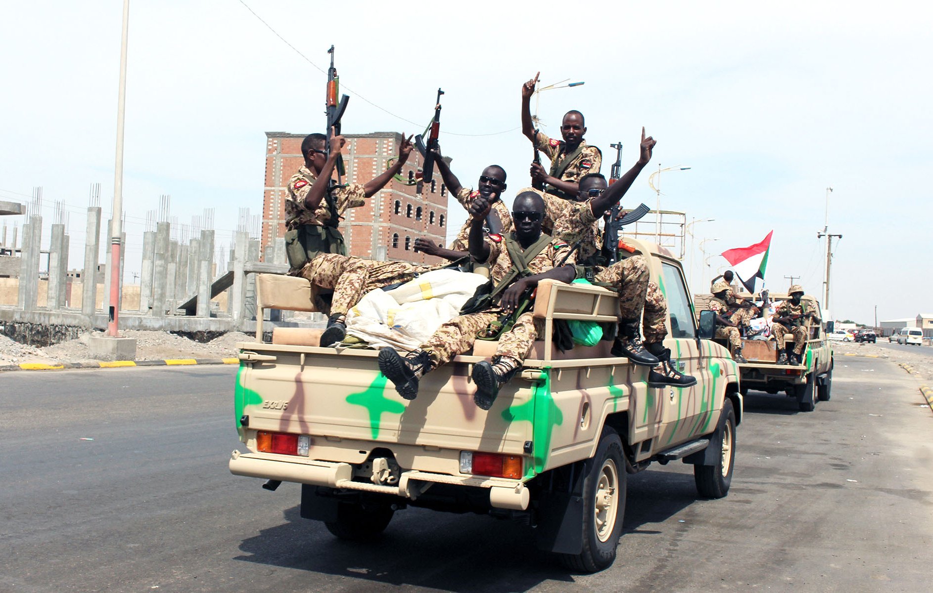 L'image montre un groupe de soldats assis sur le plateau d'un véhicule militaire. Ils portent des uniformes camouflage et tiennent des armes. Les soldats affichent des gestes de victoire en levant un doigt. En arrière-plan, on peut voir un paysage urbain inachevé avec des bâtiments en construction. L'atmosphère semble être celle d'une parade ou d'un mouvement militaire dans une zone urbaine.