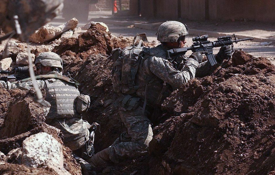 L'image montre des soldats en engagement militaire, se cachant derrière des tranchées. Ils portent des uniformes militaires et des casques, et sont armés. Le terrain est marqué par des gravats et de la terre, suggérant un environnement de combat. L'atmosphère est tendue, montrant la préparation et la vigilance des soldats face à un danger.
