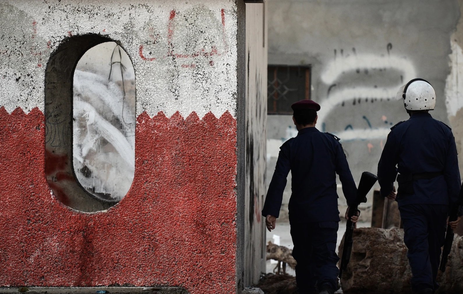 تظهر في الصورة جداراً مُرسّماً بلونين، أحمر وأبيض، مع وجود فتحة مستديرة في الجدار. يسير شخصان يرتديان زيّ قوات الأمن، أحدهما يحمل سلاحاً. الخلفية تُظهر جدراناً مُتضرّرة وكتابات على الجدران، مما يوحي بأجواء من التوتر أو الاحتجاجات. الصورة تعكس مشهداً حضرياً يعكس ربما حالة معينة في المنطقة.