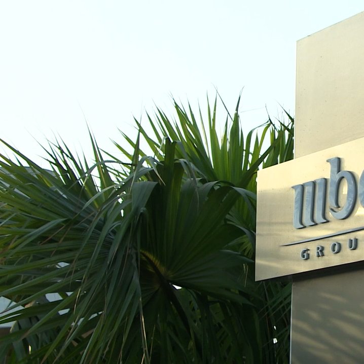 تظهر الصورة لافتة تحمل شعار "مجموعة MBC"، وهو معدني وأنيق، ويقع ضمن بيئة نباتية تتضمن أوراق نخيل وشجيرات خضراء. يبدو أن الصورة التقطت في النهار، حيث يظهر ضوء الشمس بشكل جيد. تعكس الألوان والمواد المستخدمة في التصميم الاحترافية والرقي.