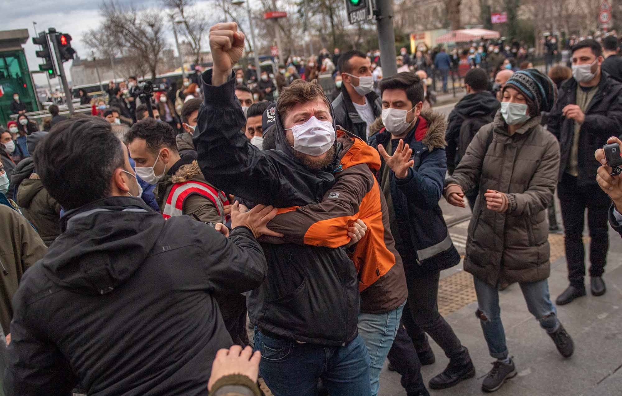 The image depicts a scene of confrontation during a protest or demonstration. Several individuals are gathered, with some appearing to engage in a physical altercation. One person, wearing a black jacket and a mask, is raising a fist, indicating a strong emotional response. People around them seem to be trying to intervene or separate those involved in the conflict. The setting appears to be an urban area, likely near a traffic light, with a backdrop of buildings and other demonstrators. The atmosphere seems tense and charged.