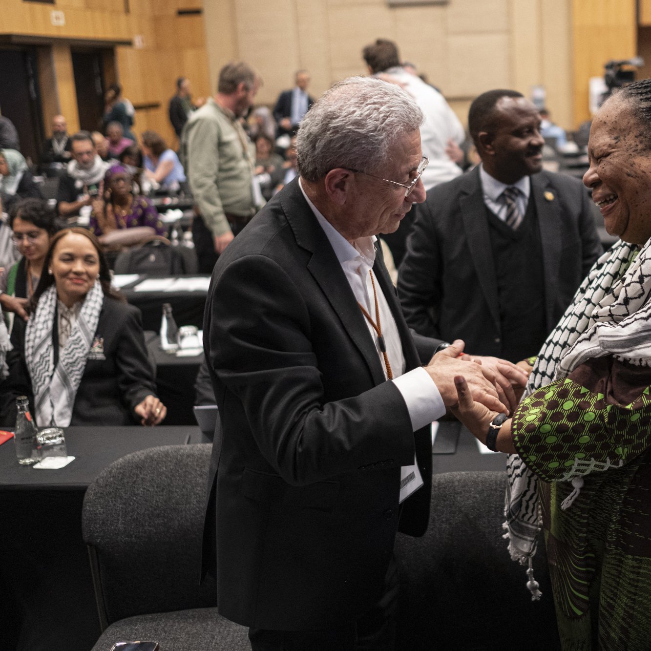 The image depicts a moment of interaction between two individuals at a conference or event. Both people are smiling and engaged in a handshake, suggesting a friendly and positive exchange. The person on the left is wearing a suit, while the individual on the right is dressed in traditional or cultural attire with a colorful shawl. In the background, there are other attendees, some seated and others standing, indicating a busy atmosphere typical of a conference. The setting appears to be indoors, likely within a large hall or meeting room.
