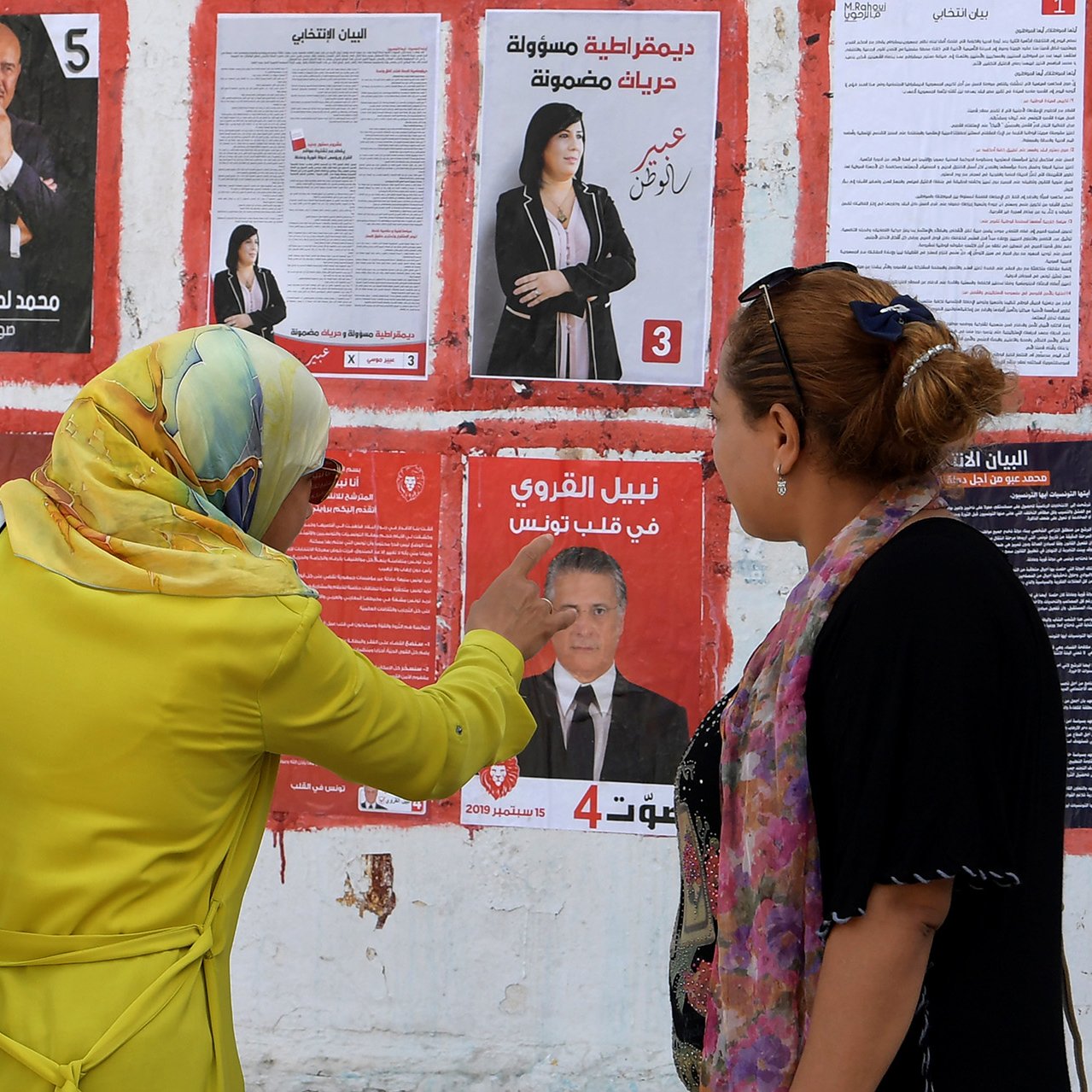 The image depicts two women standing in front of a wall covered with election posters. One woman, wearing a yellow jacket and a headscarf, appears to be pointing at one of the posters, while the other woman, dressed in a black outfit with a scarf, looks on attentively. The posters feature various candidates, showcasing their faces and names, indicative of an election campaign. The setting suggests a public space where voters might consider their options.
