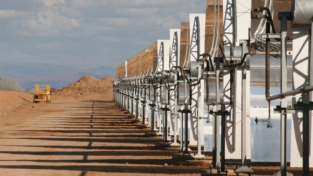 تظهر الصورة مجموعة من الألواح الشمسية المرتبطة بهياكل معدنية، تقع في منطقة صحراوية أو شبه صحراوية. الألواح مرتبة بشكل منتظم وموجهة نحو السماء، مما يشير إلى أنه يجري استغلال الطاقة الشمسية. في الخلفية، يمكن رؤية تلال ترابية وبعض السحب في السماء، مما يعطي انطباعًا عن مساحة واسعة ومفتوحة. تُظهر الصورة أيضًا طريقًا يسير فيه مركبة كبيرة تشير إلى وجود نشاط صناعي أو زراعي في المنطقة.