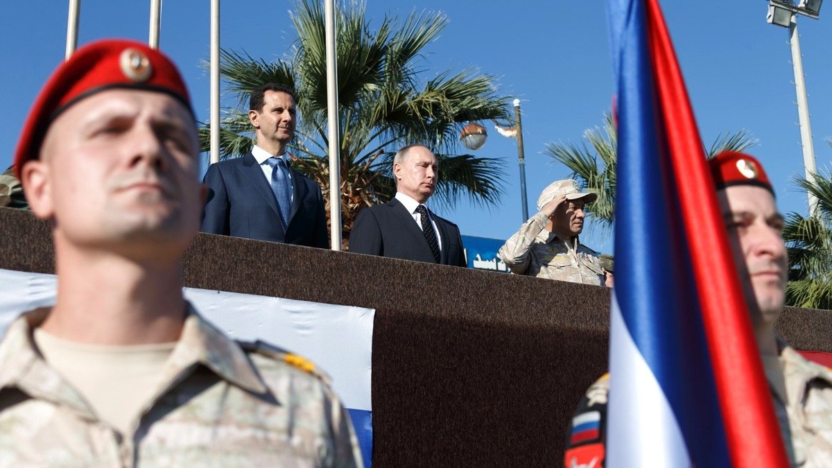 The image depicts a military ceremony or event, featuring several individuals of importance. In the foreground, soldiers in military uniforms with red berets are standing at attention. In the background, a group of officials is present, including two prominent figures positioned together: one in a suit and tie and the other in military attire. There's a large flag in the foreground, possibly representing a nation involved in the event. The setting appears outdoors, with palm trees visible, suggesting a warm climate. The overall atmosphere seems formal and significant, likely indicative of a diplomatic or military alliance.
