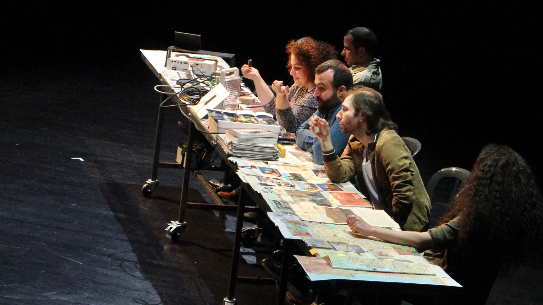 L'image montre un groupe de personnes assises autour d'une grande table sur scène, avec des ordinateurs portables et des documents éparpillés. En arrière-plan, un écran projette un schéma ou une carte, probablement liée à un contexte géographique ou politique, mentionnant des lieux comme Idleb et d'autres noms. Les participants semblent engagés dans une discussion ou une activité collaborative, ce qui suggère un travail de groupe intense. L'ambiance est dynamique, mettant en avant l'importance de l'interaction et de l'échange d'idées.