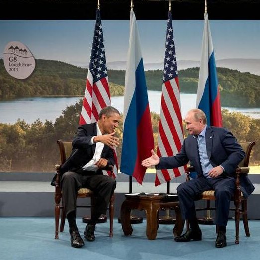 L'image montre deux dirigeants assis face à face dans un cadre formel. D'un côté, un homme souriant, vêtu d'un costume sombre avec une chemise claire, semble tendre la main vers son interlocuteur. De l'autre, un homme aux cheveux clairs, également en costume, répond avec un sourire et une expression chaleureuse. Derrière eux, on peut voir des drapeaux américains et russes, et un paysage naturel en arrière-plan. L'ensemble évoque une rencontre diplomatique ou un sommet entre ces deux pays.