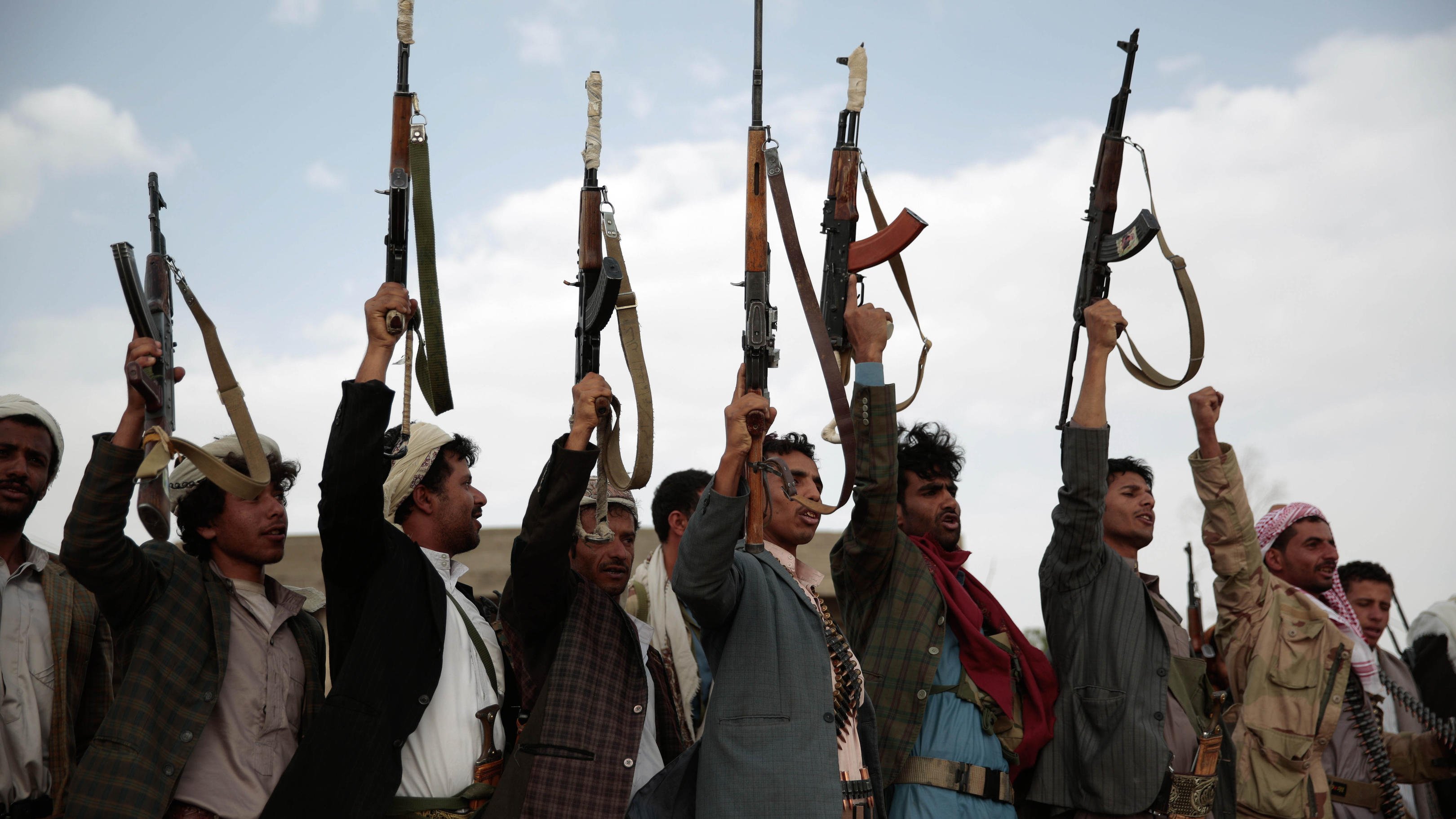 L'image montre un groupe de personnes levant des fusils en l'air. Ils semblent exprimer un sentiment de défi ou de célébration, avec des expressions déterminées sur leurs visages. Les membres du groupe portent des vêtements traditionnels et des accessoires comme des ceintures avec des munitions. Le ciel en arrière-plan est nuageux, ajoutant une atmosphère dramatique à la scène.