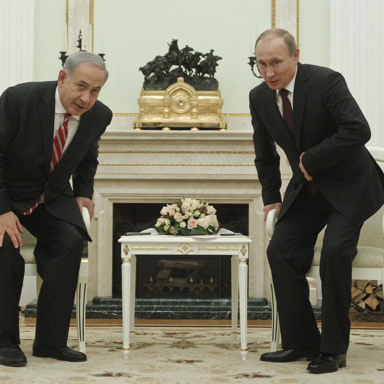 در این تصویر، دو مرد در یک اتاق رسمی نشسته‌اند. یکی از آنها لباس سیاه و کرavate قرمز بر تن دارد و به سمت جلو خم شده است. مرد دیگر نیز لباس تیره‌ای به تن دارد و در حال خم شدن به سمت اوست. در میان آن‌ها یک میز کوچک وجود دارد که روی آن گلدانی با گل‌های تازه قرار دارد. دکوراسیون اتاق بسیار کلاسیک و مجلل به نظر می‌رسد، با دیوارهای روشن و تزئینات خاص.