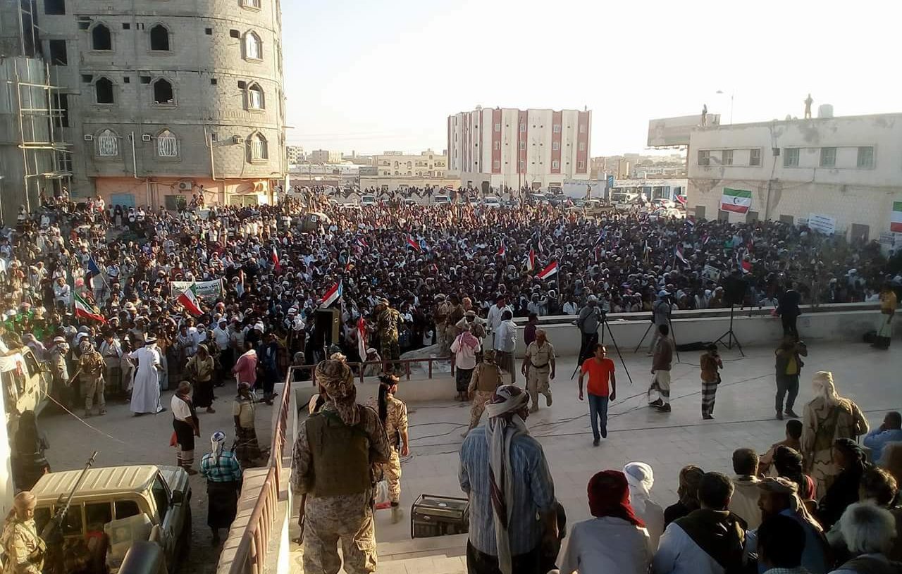 در این تصویر، یک تجمع یا راهپیمایی بزرگ در فضای باز مشاهده می‌شود. مردم زیادی در مقابل یک ساختمان قرار دارند و برخی از آن‌ها پرچم‌هایی به دست دارند. در پس‌زمینه، ساختمانی بزرگ و نیمه‌ساز دیده می‌شود، و ساختمان‌های دیگری نیز در دوردست‌ها وجود دارد. نور خورشید به صورت عمودی به این منطقه می‌تابد و جو عمومی نشان‌دهنده شور و هیجان مردم است. گروهی از افراد در جلو و عده‌ای دیگر در پشت آن‌ها نشسته‌اند یا ایستاده‌اند.